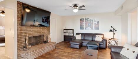 A living room with a brick fireplace, a ceiling fan, a sectional sofa, and a coffee table.