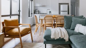 A modern kitchen with white cabinets, a coffee machine, and a refrigerator. There is a dining area with wicker chairs and a table, and a green sofa in the living room. - Close to town & hospital, quiet & private location (Sale)