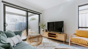 A modern living room with a large TV, a sofa, a yellow armchair, and a wicker ottoman. - Close to town & hospital, quiet & private location (Sale)