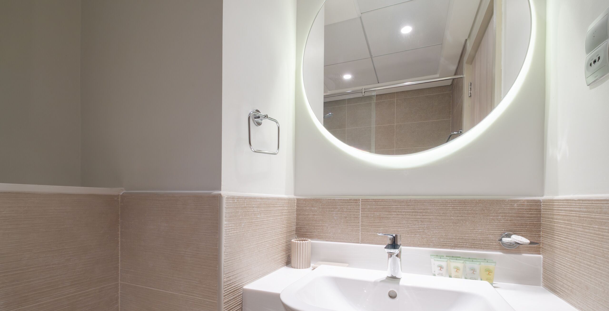 Een moderne badkamer met een wit wastafelmeubel, een spiegel met een ronde omlijsting en beige wandtegels.