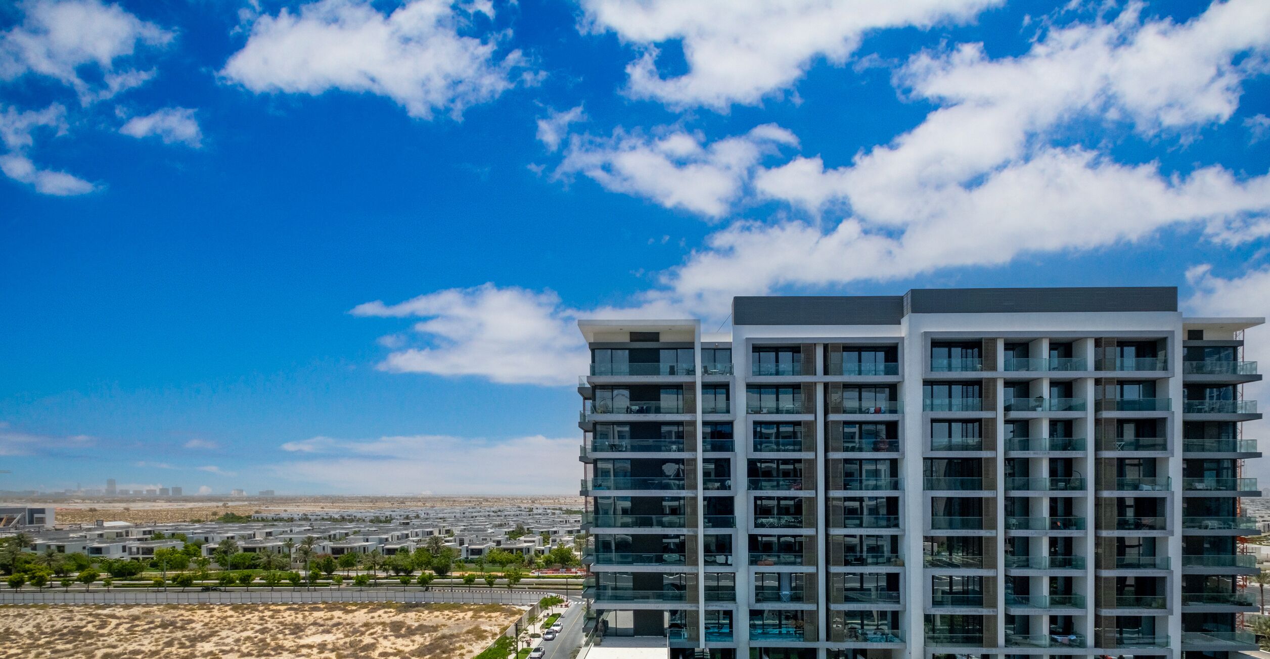 A modern high-rise building with multiple balconies, surrounded by a desert landscape and a clear blue sky.