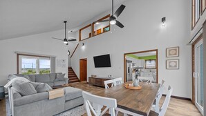 Un vaste espace de vie comprenant un canapé gris, une table à manger en bois avec des chaises blanches, un ventilateur de plafond et une cuisine visible à travers une porte ouverte.