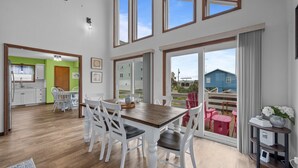Un vaste espace dédié à la salle à manger, avec une table en bois et des chaises blanches, de grandes fenêtres à cadre en bois et une vue sur une maison bleue, visible à travers la porte-fenêtre coulissante.