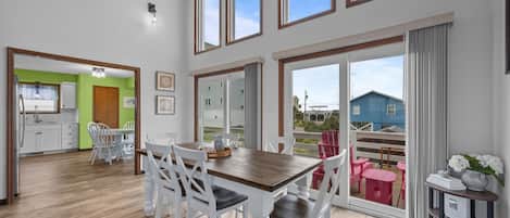 A spacious dining area with a wooden table and white chairs, large windows with wooden frames, and a view of a blue house through the sliding glass door.