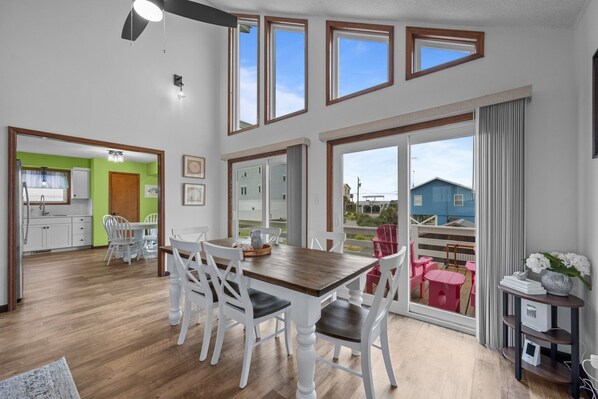 A spacious dining area with a wooden table and white chairs, large windows with wooden frames, and a view of a blue house through the sliding glass door.