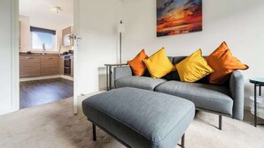 Un salon moderne avec un canapé et un pouf gris, un tableau accroché au mur et une vue sur la cuisine.