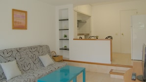 Sala de estar con sofá, una mesa de centro, televisor y una cocina visible a través de una puerta abierta.