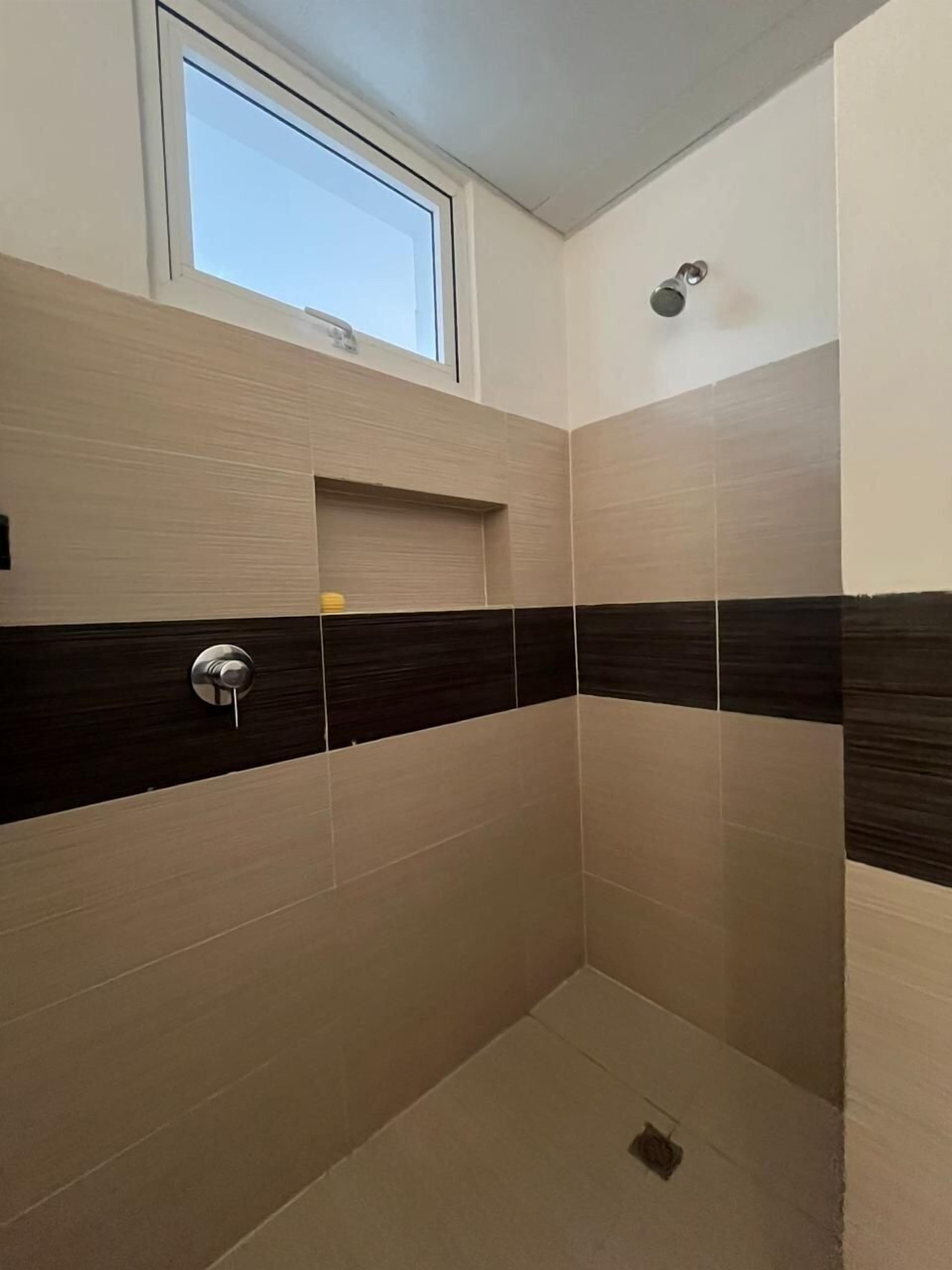 A shower with beige and dark brown tiles, a window above, and a small shelf with a yellow object.