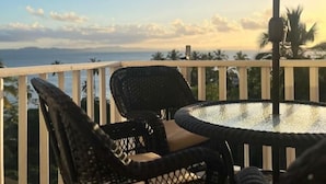 Un balcon avec une table ronde et deux fauteuils en osier, offrant une vue panoramique sur l’océan et les palmiers.