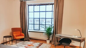 Een slaapkamer met een bed, een oranje fauteuil, een bureau met stoel, een radiator, een raam met gordijnen en een kamerplant.