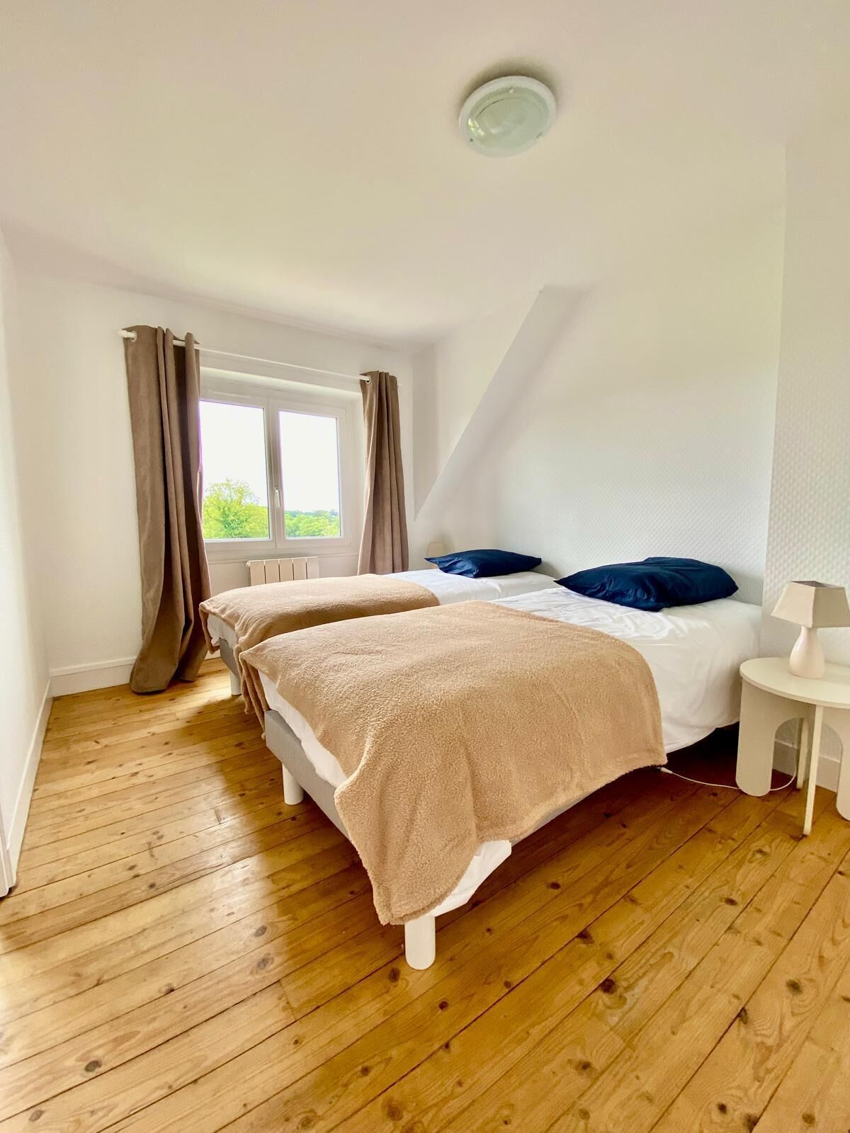 A bedroom with two beds, a window with curtains, a lamp, and wooden flooring.