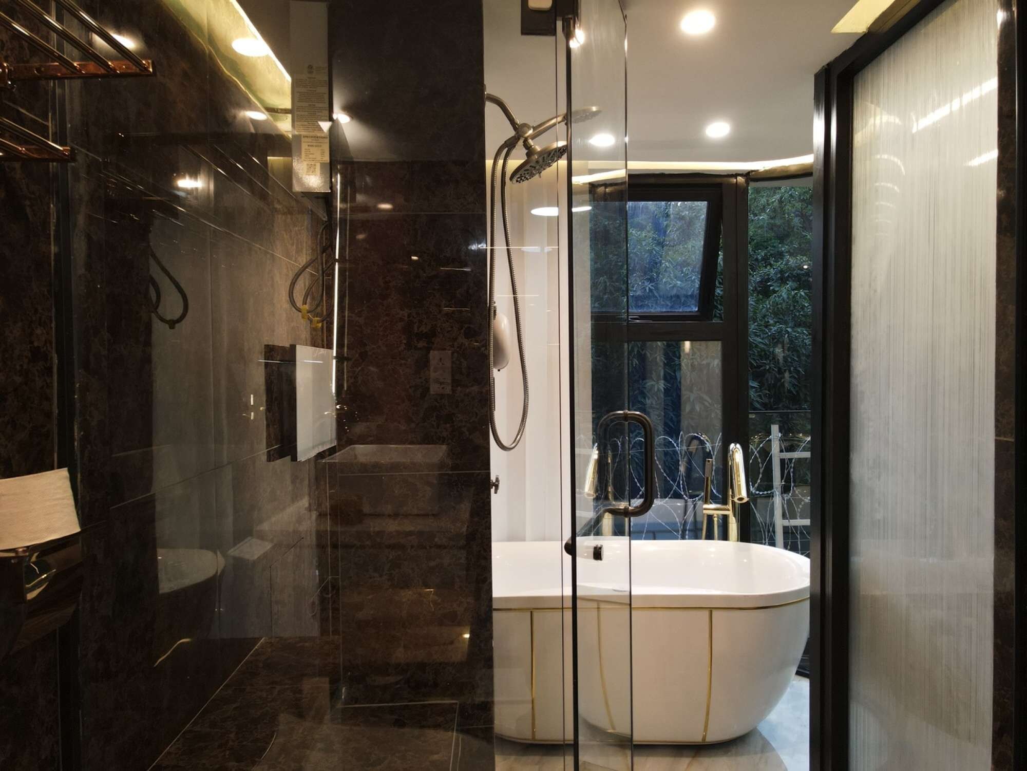 A modern bathroom with a glass-enclosed shower, a freestanding bathtub, and a window providing a view of greenery.