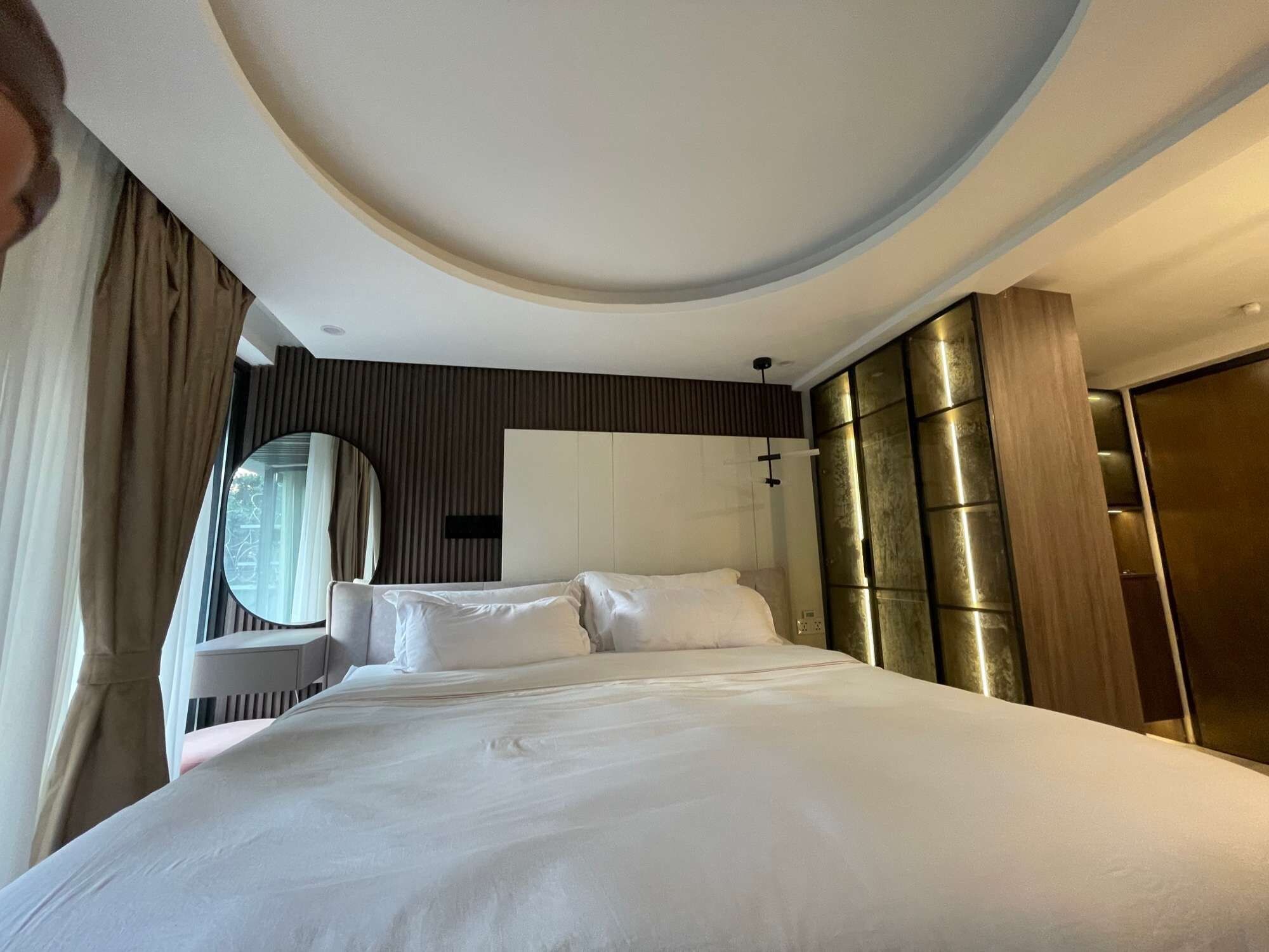 A modern hotel room with a large bed, a round mirror, a wardrobe, and a window with curtains.