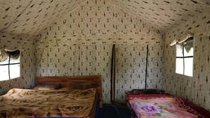 A room with a patterned wallpaper, a wooden bed with a brown blanket, a smaller bed with a floral bedspread, and a window with curtains. - The Little Pahadi (Ukhimath)
