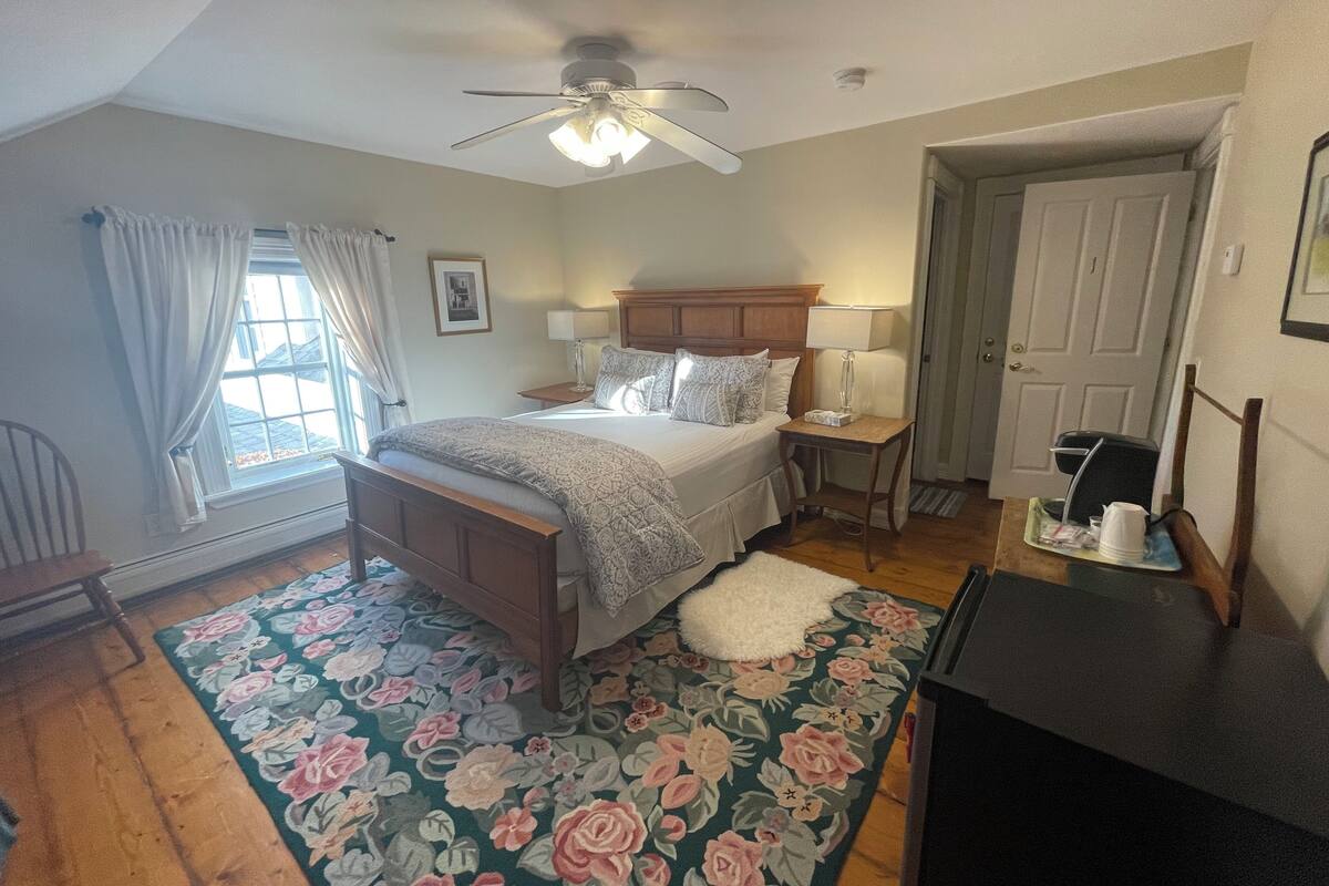 Une chambre à coucher comprenant un lit, une table de chevet, un bureau, une chaise, un ventilateur de plafond et une porte.