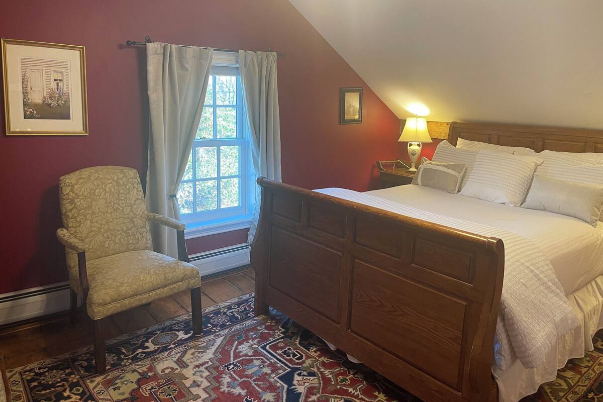 Une chambre à coucher avec un lit en bois, une chaise, une fenêtre avec des rideaux, un tapis et une lampe.