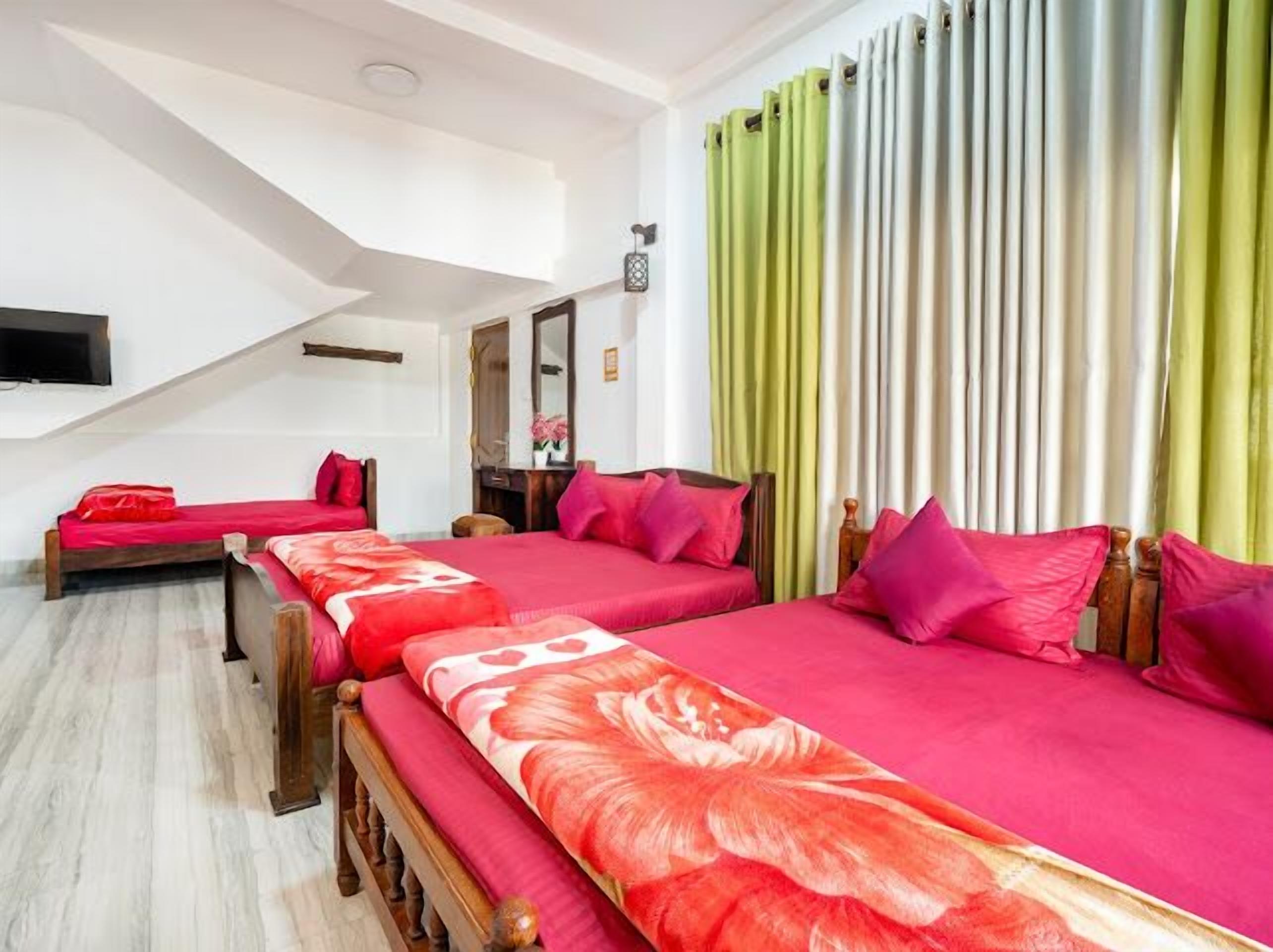 Ein Hotelzimmer mit zwei Betten, jeweils mit roter Bettwäsche und floralen Mustern, einem Holzkopfende, einem an der Wand montierten Flachbildfernseher und einem Fenster mit grünen Vorhängen.