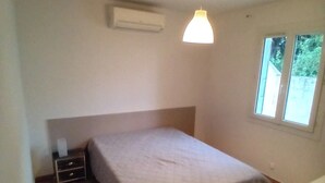 Un dormitorio pequeño y sencillo con cama, ventana y equipo de aire acondicionado.