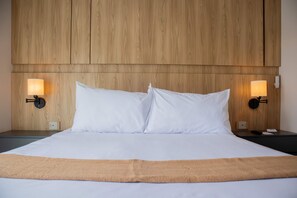 A neatly made bed with white linens and a beige throw blanket, flanked by two bedside tables with lamps and a wooden headboard.