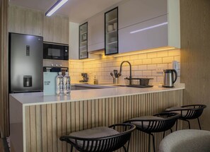 A modern kitchen with a stainless steel refrigerator, microwave, and coffee maker. There are two bottles of water on the counter and a coffee pot. The kitchen has a white countertop and a wooden backsplash.