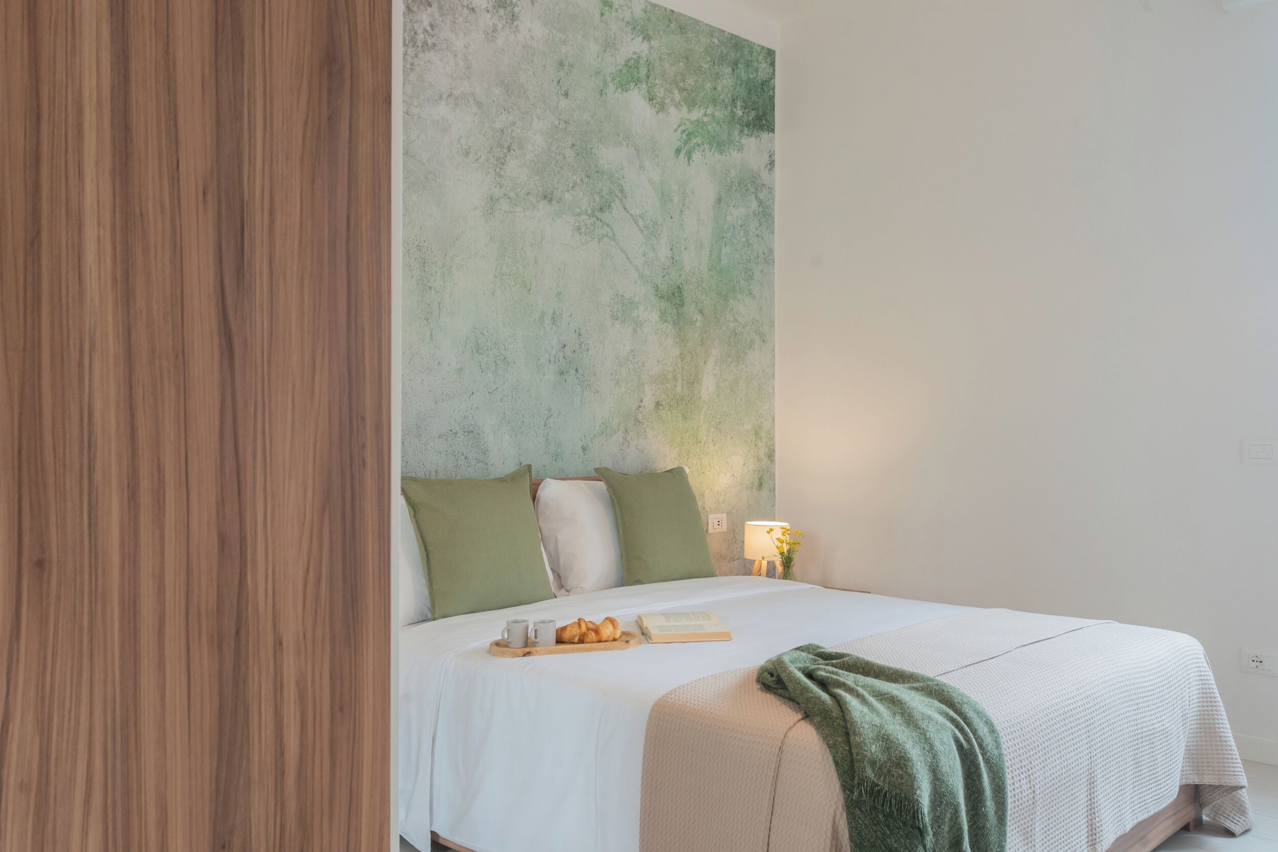 A neatly made bed with a tray of croissants and a book, a green throw blanket, and a wall with a textured green and white abstract design.