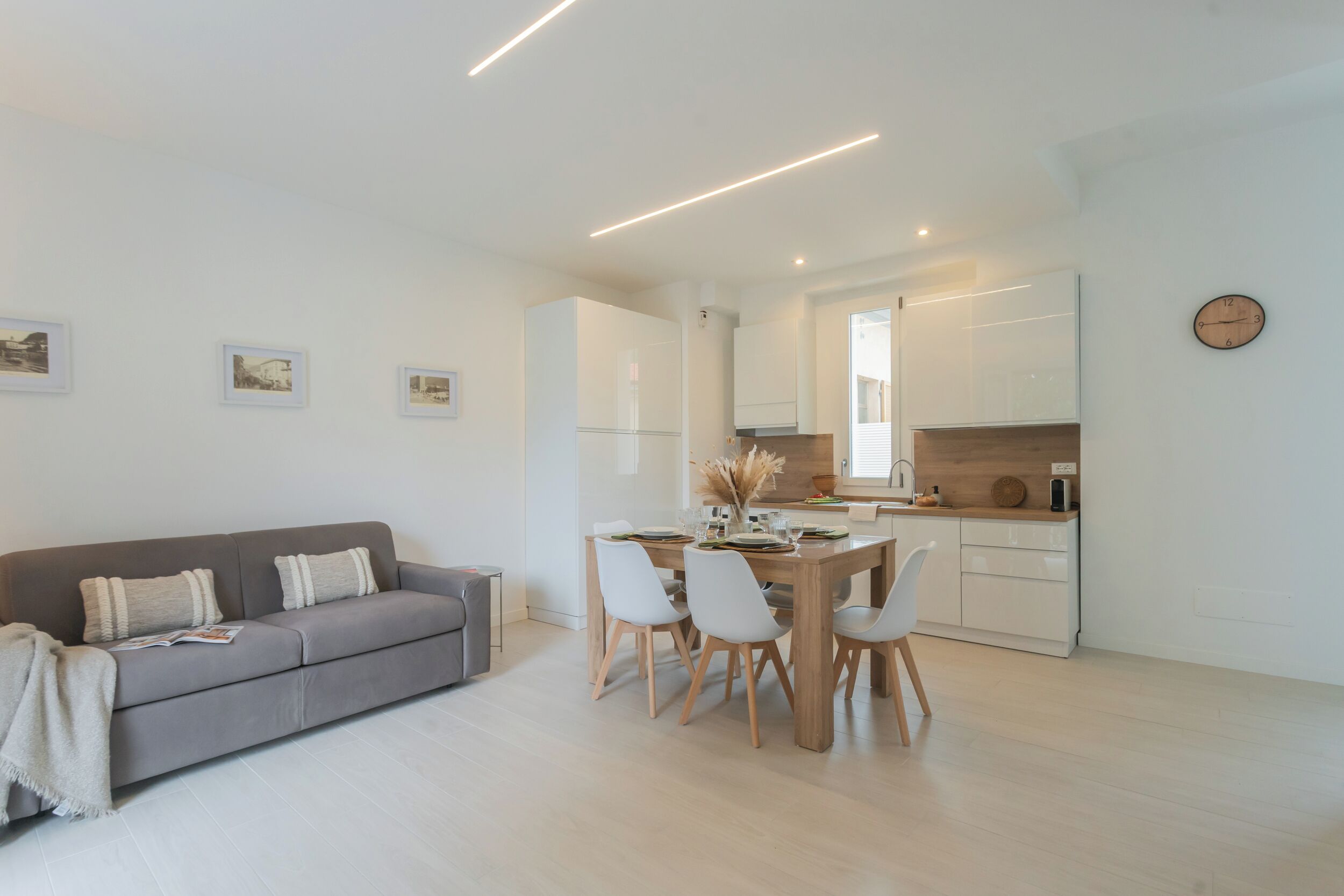 A modern living room with a grey sofa, a dining table with chairs, and a kitchen area with white cabinets and a wooden countertop.