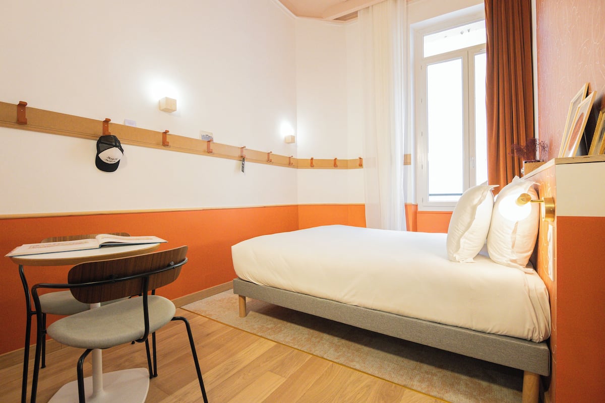 Ein Hotelzimmer mit Bett, Schreibtisch mit Stuhl, Fenster mit Vorhängen und einer Wandablage mit Kappe und einem kleinen Gegenstand.