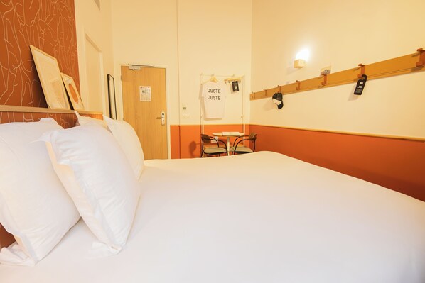 A hotel room with a bed, a desk, and a chair. There is a coat rack with hats and a white t-shirt hanging on it.