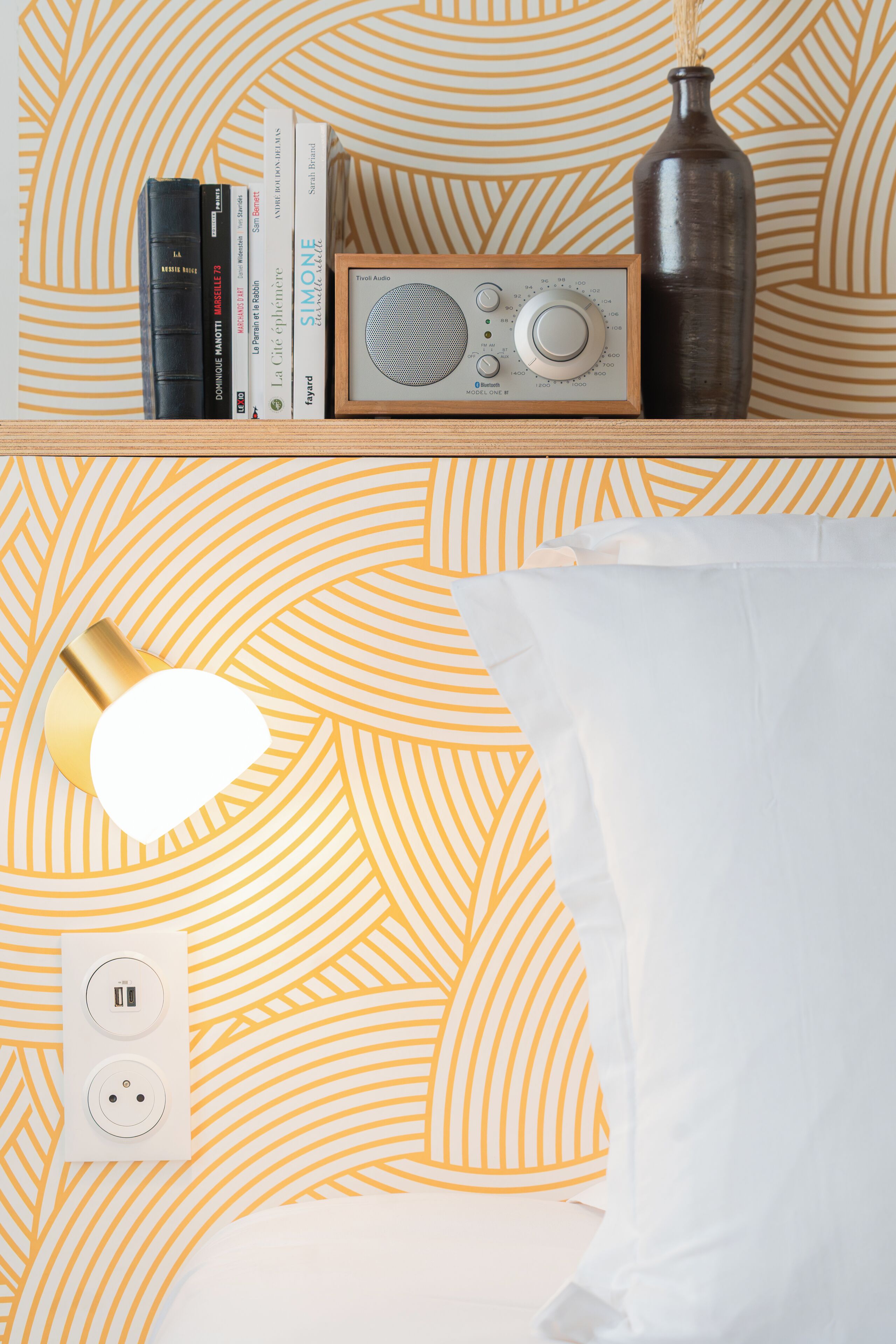 Ein ordentlich eingerichteter Nachttisch mit Radio, Büchern, einer Vase und einer Lampe auf einer gestreiften Tagesdecke.