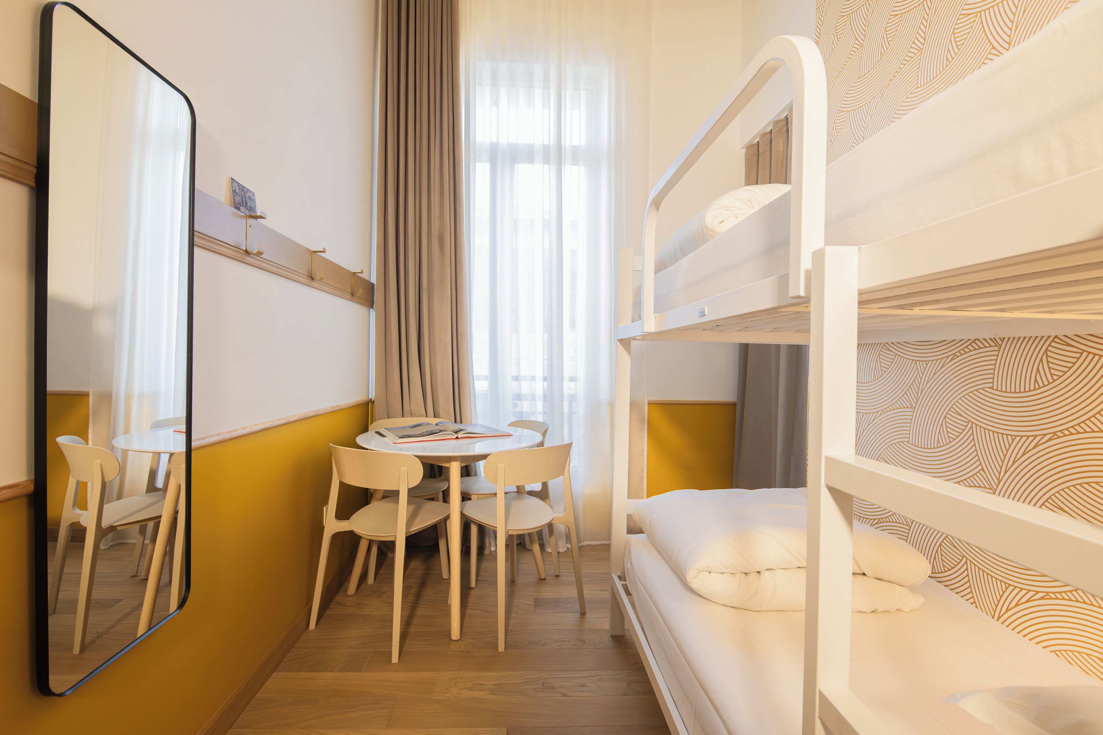 Une chambre d’hôtel avec un lit superposé, une table ronde avec des chaises, un miroir et une fenêtre avec des rideaux.
