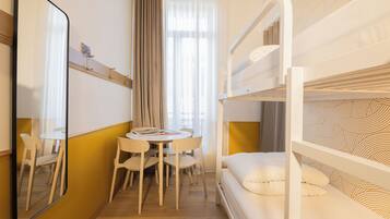 Une chambre d’hôtel avec un lit superposé, une table ronde avec des chaises, un miroir et une fenêtre avec des rideaux.