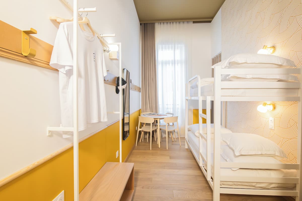 Ein Hotelzimmer mit Etagenbetten, einem kleinen Tisch mit Stühlen, einer Bank, einem Spiegel und einem Fenster mit Vorhängen.
