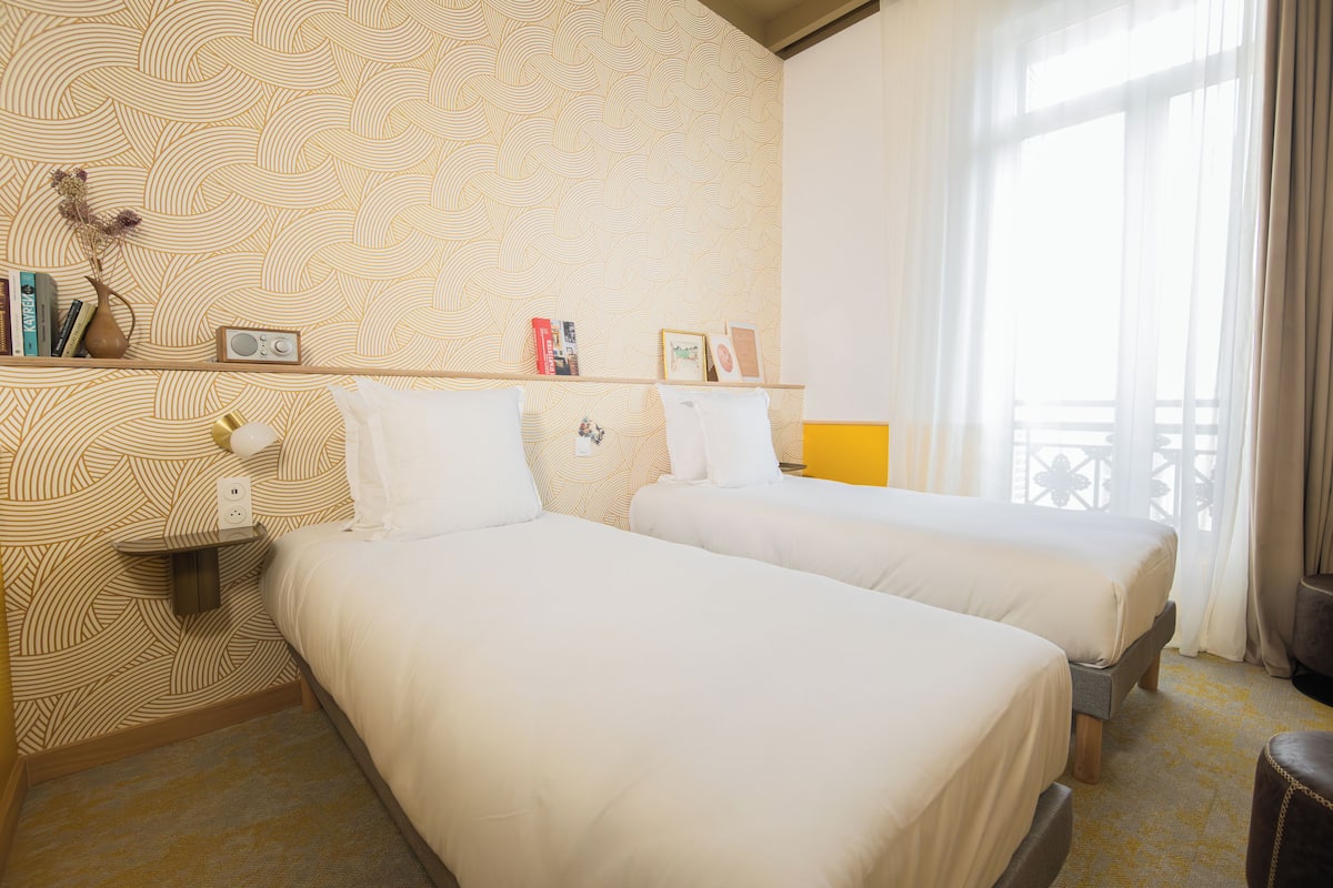 Ein Hotelzimmer mit zwei Betten, einem Regal mit Büchern und einem Radio sowie einem Fenster mit Vorhängen.