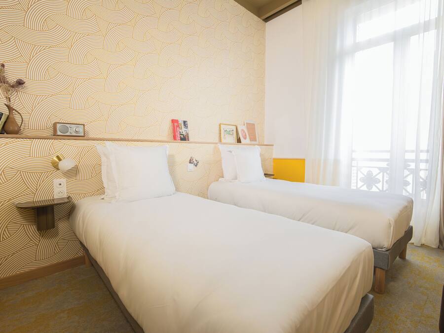 Quarto de hotel com duas camas, uma estante com livros e um rádio, e uma janela com cortinas.