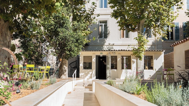 A courtyard with a pathway leading to a building entrance, surrounded by greenery and outdoor seating. - Grand Juste (Marseille)