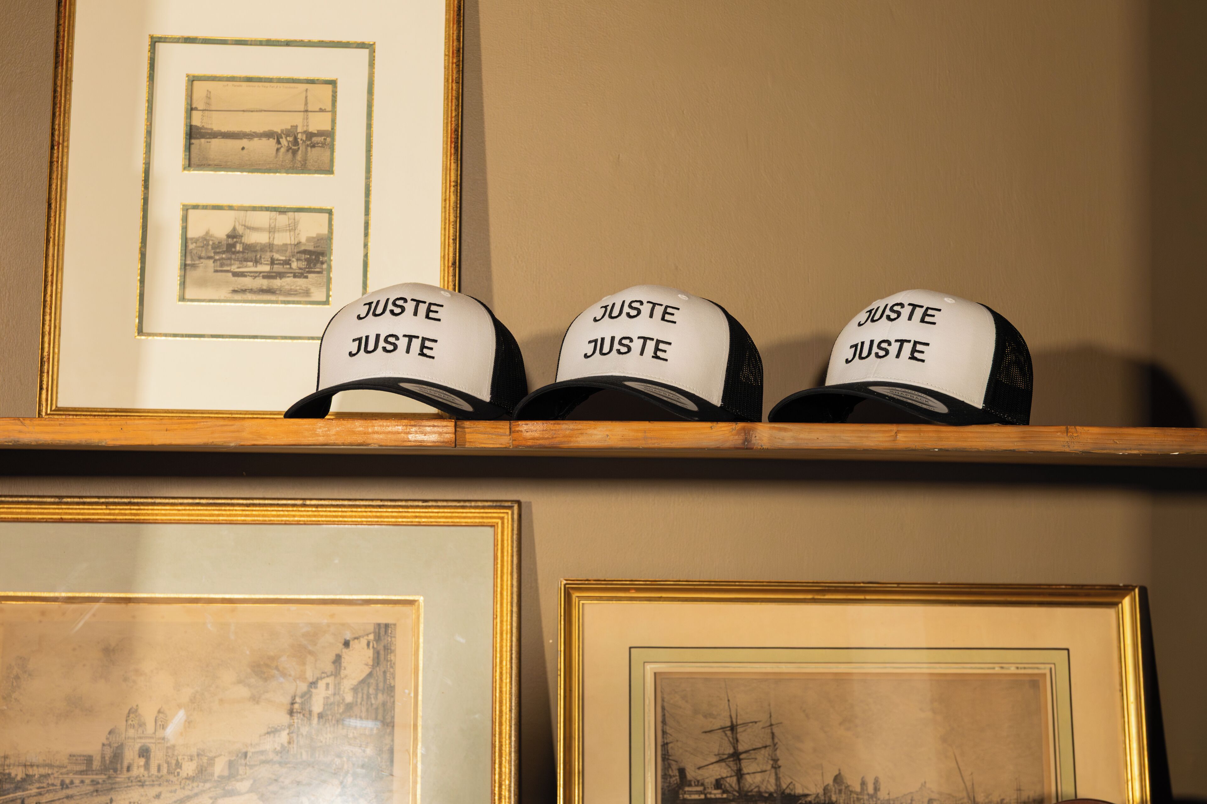 Three caps with the word "JUSTE" on a shelf above framed pictures.