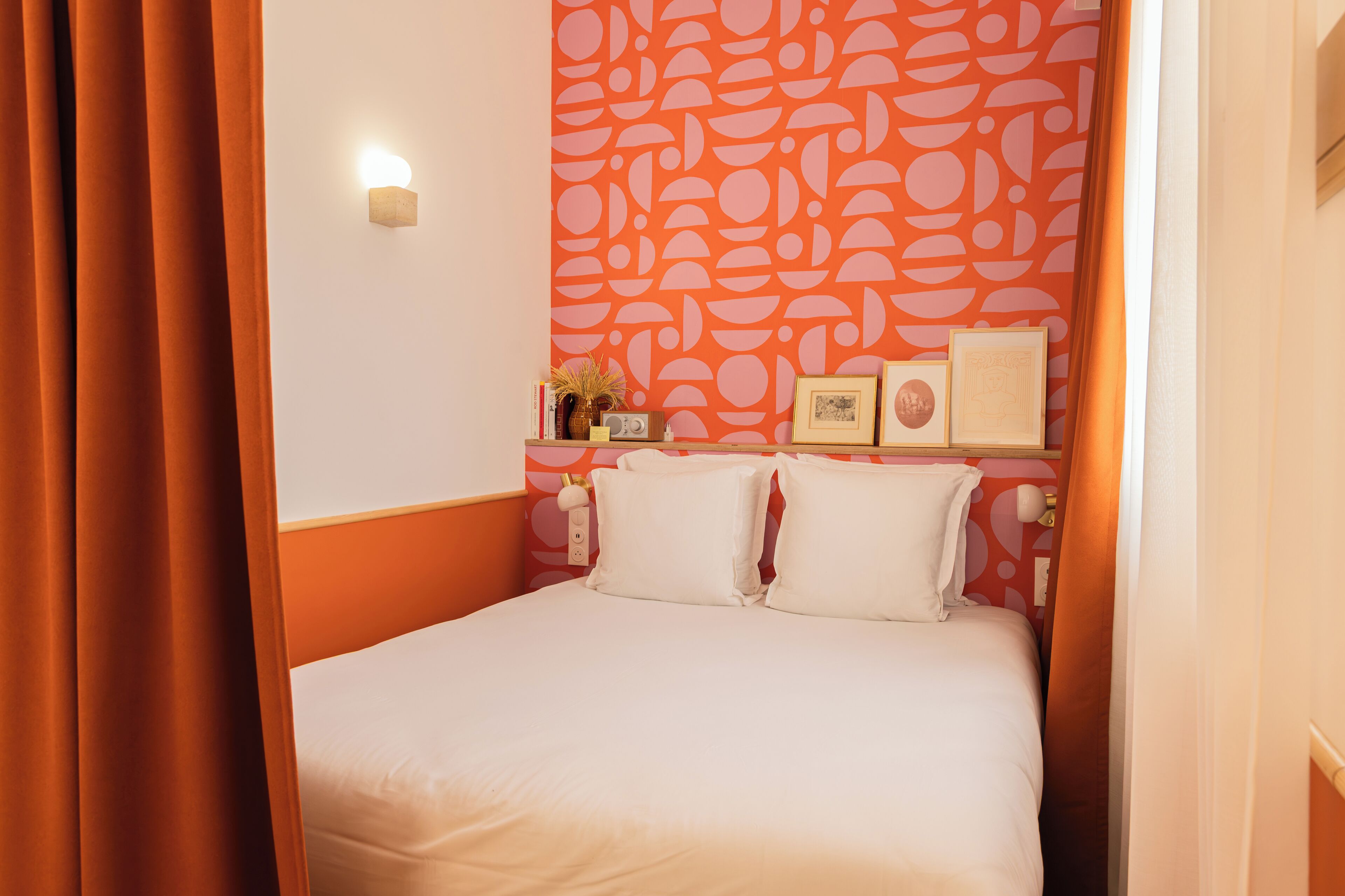 Ein ordentlich bezogenes Bett mit weißen Bettwäsche, eine Wand mit einem geometrischen Orange-Muster und ein Nachttisch mit einem Radio und gerahmten Bildern.
