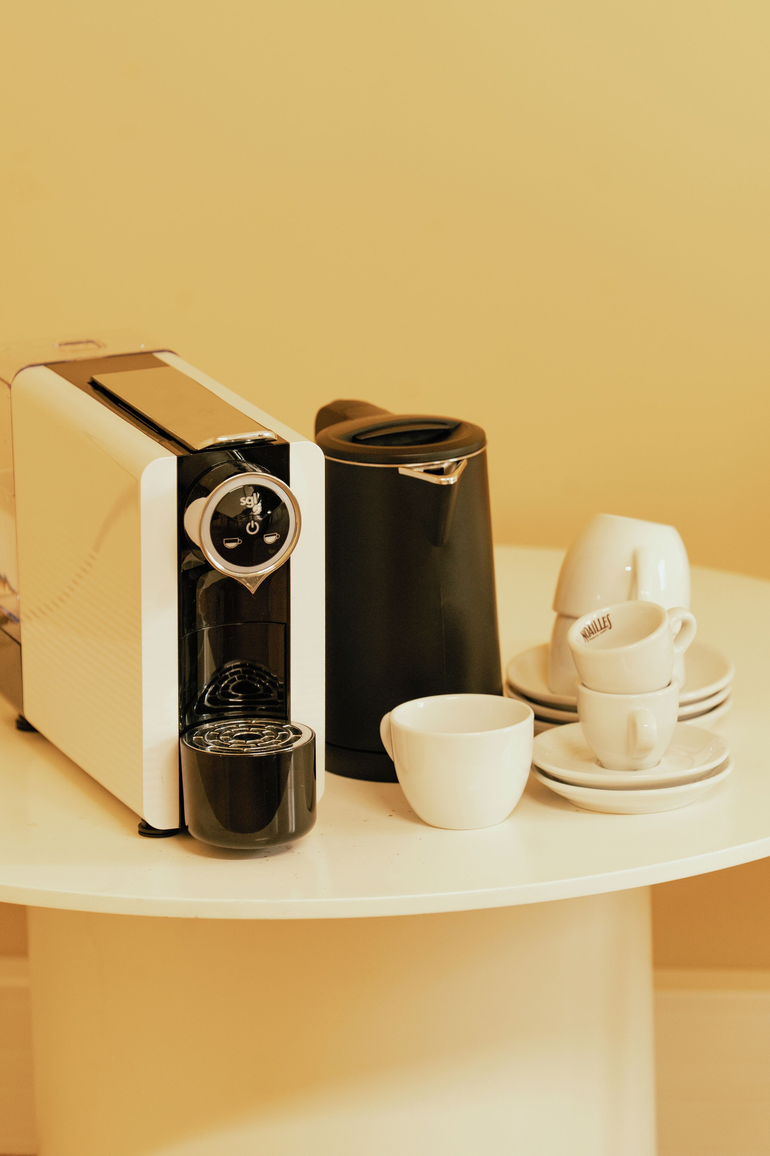 A coffee maker, a black thermos, and white cups and saucers on a white table.