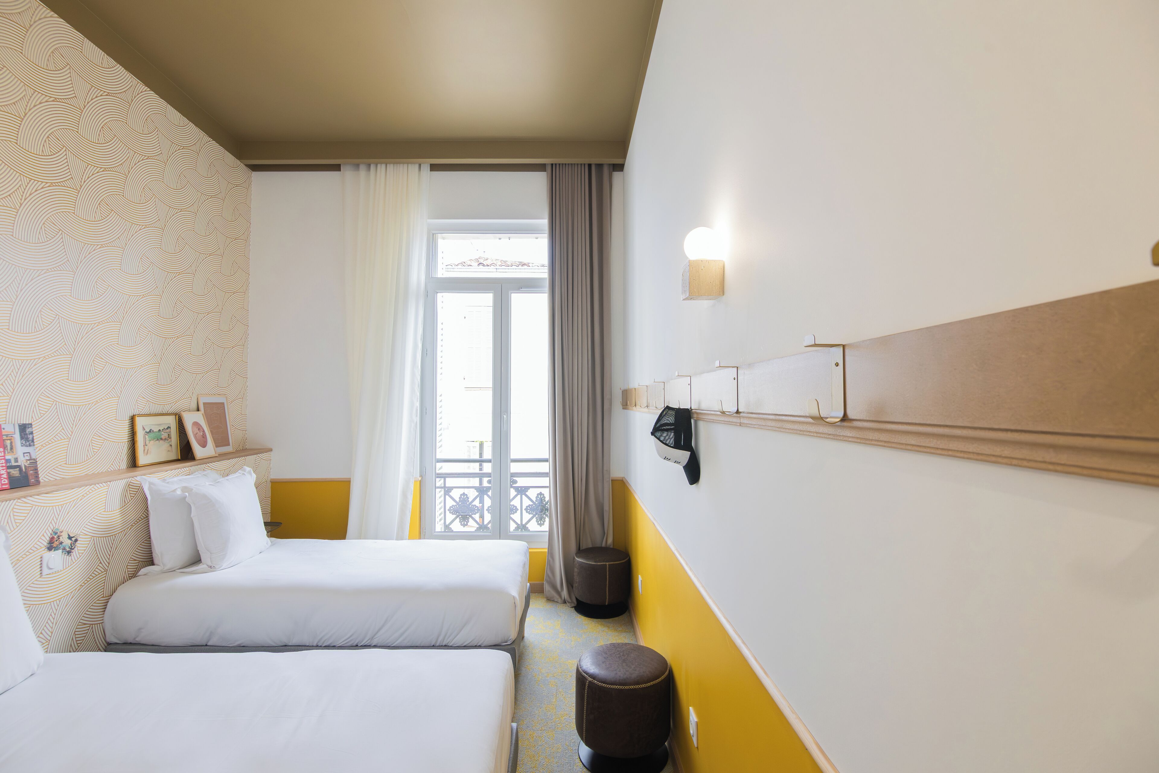 A hotel room with two beds, a window with curtains, a wall-mounted shelf with pictures, and a small stool.