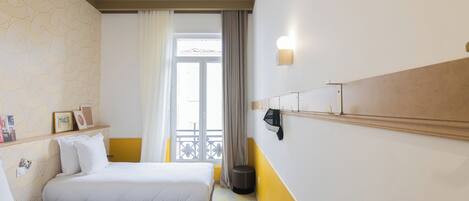 A hotel room with two beds, a window with curtains, a wall-mounted shelf with pictures, and a small stool.