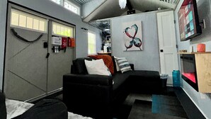 Un espace de vie compact avec un canapé noir, un mur blanc orné d’une œuvre d’art en forme de cœur, un ventilateur de plafond et un téléviseur fixé au mur.