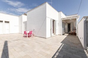 Un edificio moderno con techo plano, un patio cubierto con mesa y sillas, y un cielo despejado.