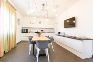 A modern kitchen with white cabinets, a dining table with chairs, a TV mounted on the wall, and a chandelier.