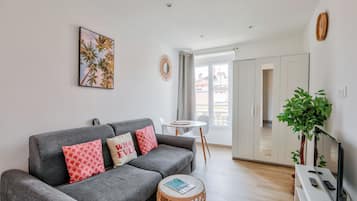 A modern living room with a grey sofa, a small round table, a TV stand, and a large window with a view of buildings.