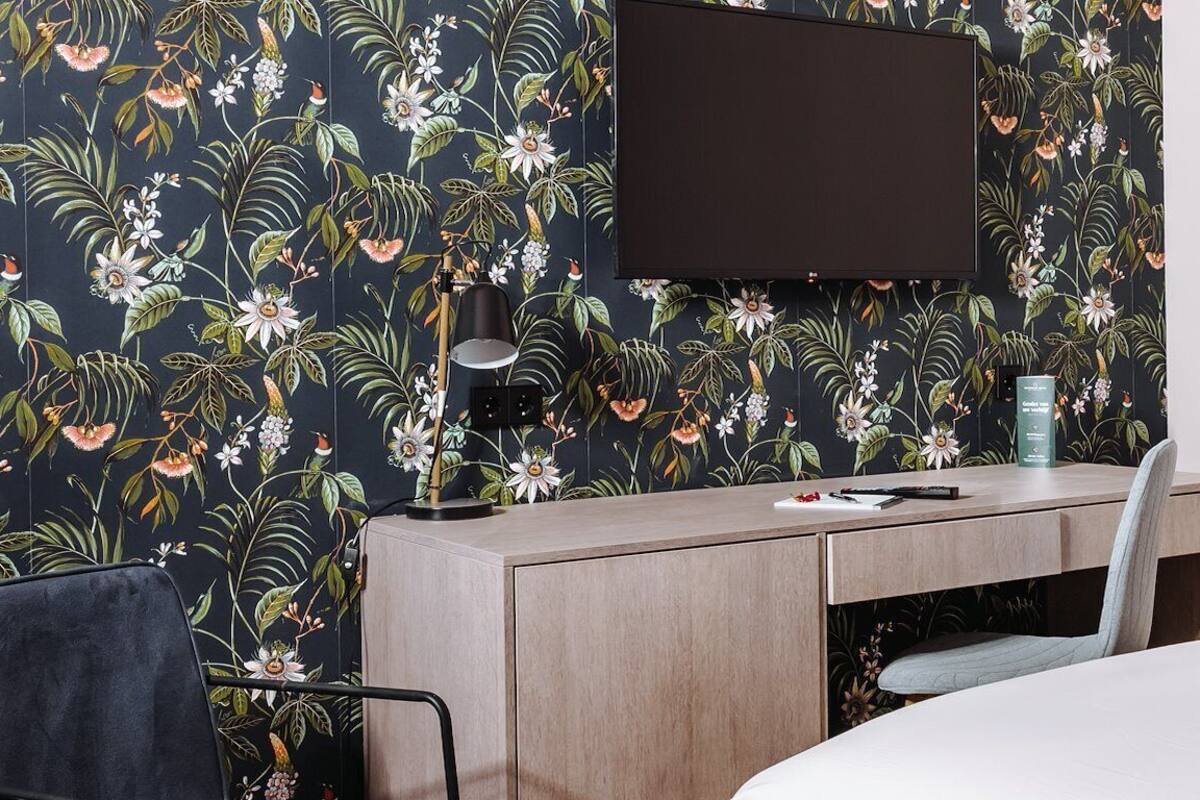 Habitación de hotel con un escritorio de madera, una silla, una cama, un televisor de pantalla plana montado en la pared y un papel tapiz con motivos florales.