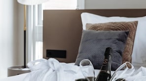 Una cama bien hecha con una botella de champán y copas, una lámpara de noche y una ventana con cortinas.