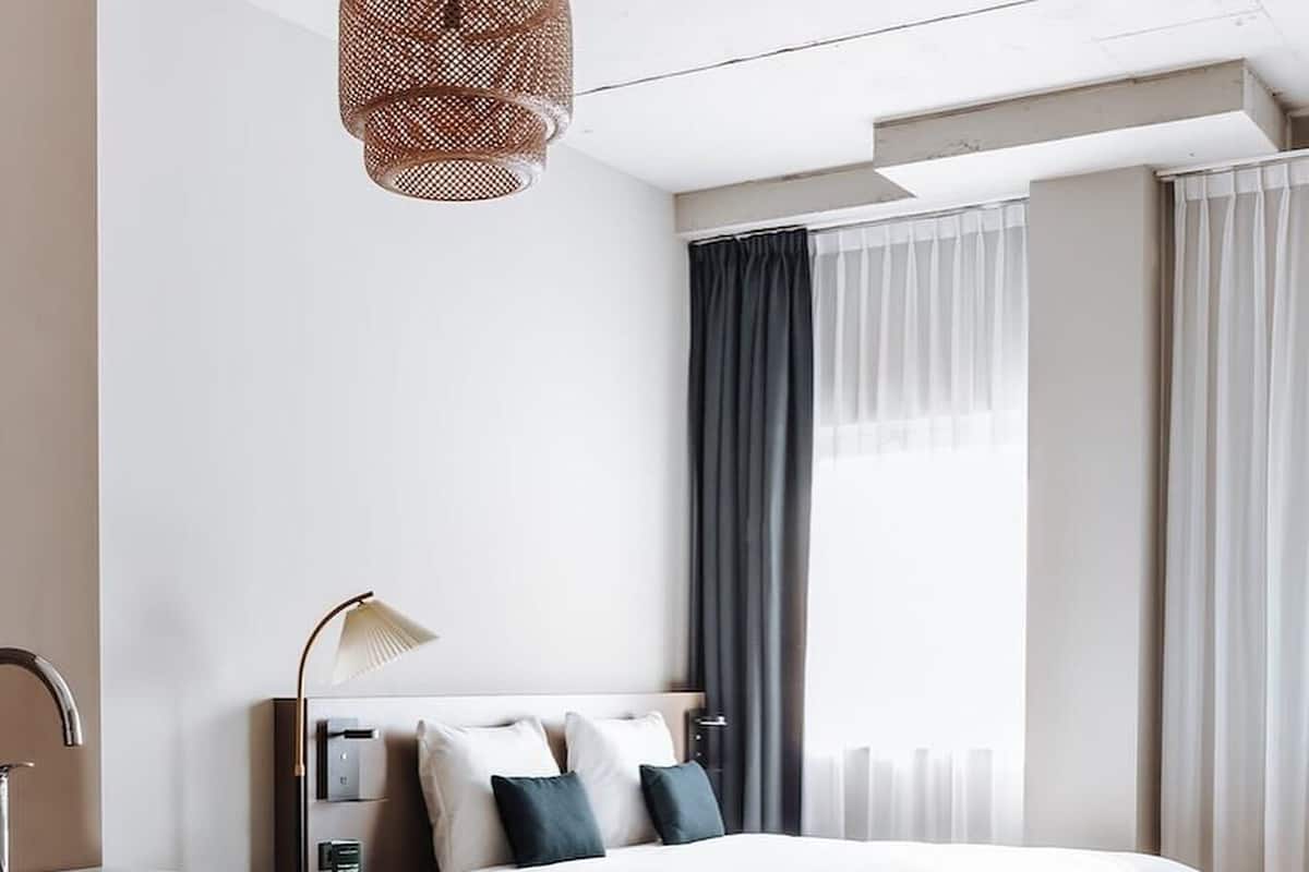 Una habitación de hotel moderna con cama, escritorio con lámpara, ventana con cortinas y piso de madera.