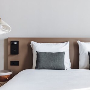 Una cama bien tendida con almohadas blancas y grises, un cabecero moderno y una mesita de noche con lámpara.