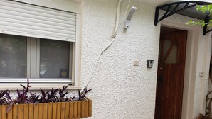 Een witte, gestructureerde muur met een houten plantenbak met planten met paarse bladeren, een raam met zonwering, een deur met houten kozijn en een potplant aan de rechterkant.