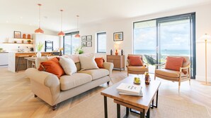 A modern living room with a sofa, armchairs, and a dining table. There is a kitchen area with a dining table and chairs. The room has large windows with a view of the ocean. - The Lighthouse is an end of terrace four bedroomed seafront retreat with a hot tub and incredible vi (Widemouth Bay)